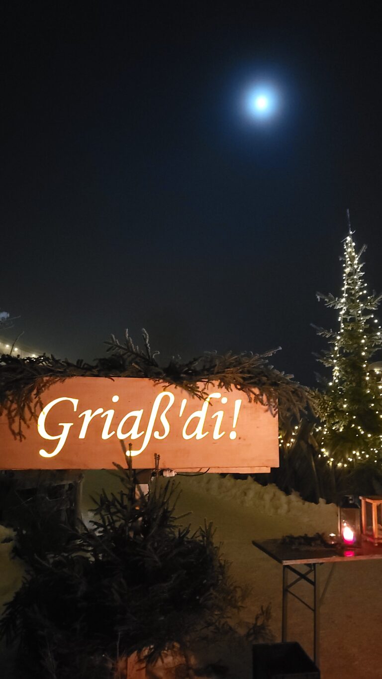 Leuchtendes Griaß'di Holzschild mit Tannenzweigen geschmückt als herzliche Begrüßung zur Weihnachtsroas.